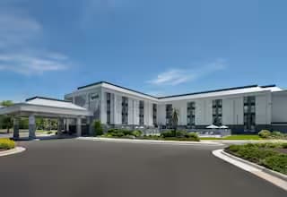 Modern white and grey multi-story hotel with a covered entrance and landscaping under blue skies.