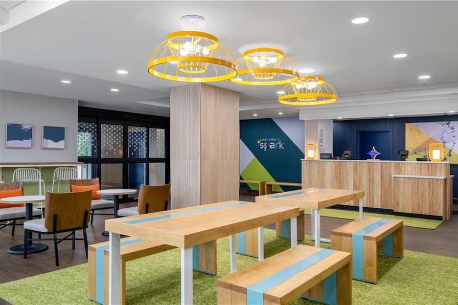 Modern hotel lobby featuring yellow cage pendant lights, wooden communal tables, and a reception desk.